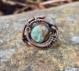 This handcrafted ring features a vibrant African green opal wrapped in warm copper. The stone’s natural patterns and earthy green tones are enhanced by the copper setting, creating a bold yet elegant design. Sized at 6, this ring is a unique piece that carries both natural beauty and artisan craftsmanship.