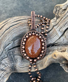 This eye-catching necklace features a shimmering Goldstone centerpiece, known for its rich, warm hue and subtle sparkle. Wrapped in handcrafted copper wire and surrounded by copper beads, the setting enhances the stone's radiant glow while adding a rustic, artisanal charm. A striking piece that blends elegance with earthy warmth.