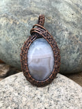 This soothing pendant features a soft, sky-blue Lace Agate stone with delicate natural banding, wrapped in warm copper wire to complement its calming tones. The handcrafted wirework enhances the stone’s serene beauty.