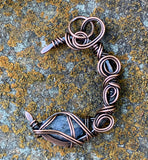 This Heavy Gauge Hammered Copper Moon Pendant is a handcrafted statement piece that features a rugged, organic design. The pendant is shaped into a crescent moon from heavy gauge copper, which has been hammered to create texture and depth. Adorning the moon are intricate copper swirls, adding a delicate touch to the bold copper design. The pendant is further enhanced by a Raw Tanzanite accent stone, which brings a pop of vibrant blue-violet color and metaphysical significance to the piece.