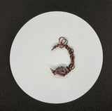 This Heavy Gauge Hammered Copper Moon Pendant is a handcrafted statement piece that features a rugged, organic design. The pendant is shaped into a crescent moon from heavy gauge copper, which has been hammered to create texture and depth. Adorning the moon are intricate copper swirls, adding a delicate touch to the bold copper design. The pendant is further enhanced by a Raw Tanzanite accent stone, which brings a pop of vibrant blue-violet color and metaphysical significance to the piece.
