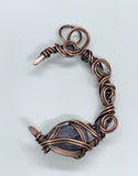 This Heavy Gauge Hammered Copper Moon Pendant is a handcrafted statement piece that features a rugged, organic design. The pendant is shaped into a crescent moon from heavy gauge copper, which has been hammered to create texture and depth. Adorning the moon are intricate copper swirls, adding a delicate touch to the bold copper design. The pendant is further enhanced by a Raw Tanzanite accent stone, which brings a pop of vibrant blue-violet color and metaphysical significance to the piece.