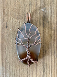 This Dark Brown to Clear Agate Tree of Life Pendant is a stunning piece of boho-inspired jewelry featuring a natural agate stone with a gradient from dark brown to clear. The pendant is beautifully wrapped in copper and showcases a detailed Tree of Life design, making it a unique and nature-inspired accessory.