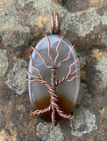 This Dark Brown to Clear Agate Tree of Life Pendant is a stunning piece of boho-inspired jewelry featuring a natural agate stone with a gradient from dark brown to clear. The pendant is beautifully wrapped in copper and showcases a detailed Tree of Life design, making it a unique and nature-inspired accessory.