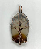 This Dark Brown to Clear Agate Tree of Life Pendant is a stunning piece of boho-inspired jewelry featuring a natural agate stone with a gradient from dark brown to clear. The pendant is beautifully wrapped in copper and showcases a detailed Tree of Life design, making it a unique and nature-inspired accessory.