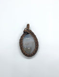 This soothing pendant features a soft, sky-blue Lace Agate stone with delicate natural banding, wrapped in warm copper wire to complement its calming tones. The handcrafted wirework enhances the stone’s serene beauty.