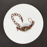 This Copper Ore Jasper Bracelet features a stunning Copper Ore Jasper stone as its focal point, surrounded by intricate, rolling hand-woven copper weaves. The bracelet combines the natural beauty of the jasper stone with the warmth and artistry of copper wirework, creating a bold and elegant accessory that showcases skilled craftsmanship. The earthy tones of the stone paired with the flowing copper design make this a striking and versatile piece.