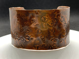 This striking wide copper cuff bracelet is hand-embossed with intricate patterns. The rich, warm tones of the copper are enhanced by the detailed embossing, creating a bold yet elegant statement piece.