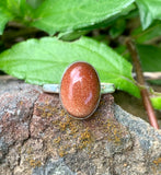 A goldstone glimmers like captured starlight, held in smooth sterling silver — this ring is radiant, refined, and full of warmth.