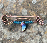 This unique, handcrafted copper pin is made from heavy gauge copper, featuring graceful copper swirls that add texture and movement to the design. The pin is adorned with vibrant blue glass beads and a dazzling rainbow glass leaf accent, bringing a touch of color and whimsy to the piece. Perfect for adding an artistic flair to your outfit, this one-of-a-kind copper pin embodies a blend of rustic elegance and playful charm.