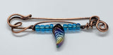 This unique, handcrafted copper pin is made from heavy gauge copper, featuring graceful copper swirls that add texture and movement to the design. The pin is adorned with vibrant blue glass beads and a dazzling rainbow glass leaf accent, bringing a touch of color and whimsy to the piece. Perfect for adding an artistic flair to your outfit, this one-of-a-kind copper pin embodies a blend of rustic elegance and playful charm.