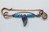 This unique, handcrafted copper pin is made from heavy gauge copper, featuring graceful copper swirls that add texture and movement to the design. The pin is adorned with vibrant blue glass beads and a dazzling rainbow glass leaf accent, bringing a touch of color and whimsy to the piece. Perfect for adding an artistic flair to your outfit, this one-of-a-kind copper pin embodies a blend of rustic elegance and playful charm.