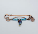 This unique, handcrafted copper pin is made from heavy gauge copper, featuring graceful copper swirls that add texture and movement to the design. The pin is adorned with vibrant blue glass beads and a dazzling rainbow glass leaf accent, bringing a touch of color and whimsy to the piece. Perfect for adding an artistic flair to your outfit, this one-of-a-kind copper pin embodies a blend of rustic elegance and playful charm.