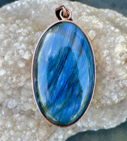 This bold pendant showcases a striking blue labradorite with vibrant flashes of color that dance in the light. Set in copper, the design enhances the stone’s natural iridescence and mystical appeal. A powerful statement piece, perfect for those drawn to the magic of labradorite.