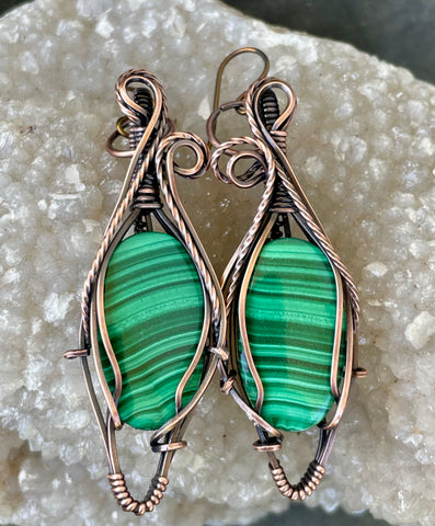 These handcrafted earrings feature rich green malachite stones wrapped in a combination of square and twisted square copper wire, adding depth and dimension to the design. The swirling bands of the malachite beautifully complement the warm copper tones, while hypoallergenic niobium ear wires ensure all-day comfort. A perfect blend of bold elegance and natural beauty—ideal for everyday wear or special occasions.