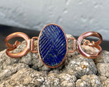 This bracelet features a richly colored carved Lapis Lazuli centerpiece, etched in a criss-cross pattern that adds eye-catching shine and texture. Set in warm copper wire, the design blends bold color with graceful form. The adjustable fit ensures comfort and versatility, making this a striking accessory for any wrist.