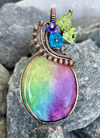 This vibrant pendant features a stunning dyed Rainbow Quartz centerpiece, showcasing a spectrum of colors that beautifully catch the light. Wrapped in warm copper, the pendant is adorned with delicate glass flowers and a charming leaf accent, adding an artistic touch to the design. This unique piece celebrates the beauty of nature and color, making it a perfect statement accessory for any outfit. Whether for everyday wear or special occasions, this pendant is sure to brighten your jewelry collection!