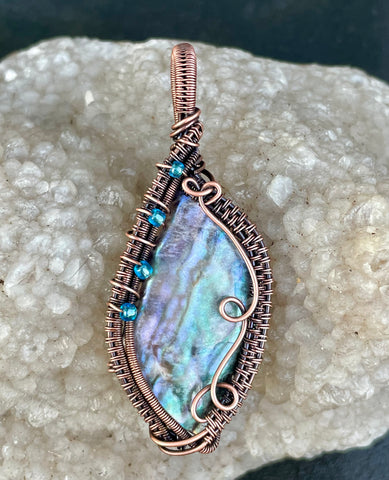 This Colorful Pen Shell Pendant features a vibrant pen shell encased in an intricate, handwoven copper wire design and swirling copper waves, complemented by delicate glass bead accents. The pen shell, known for its iridescent and colorful surface, showcases a spectrum of hues ranging from purple to flashes of blues and greens, making this pendant a true visual delight. The copper wire wrapping adds warmth and texture, while the glass beads offer a subtle touch of sparkle.