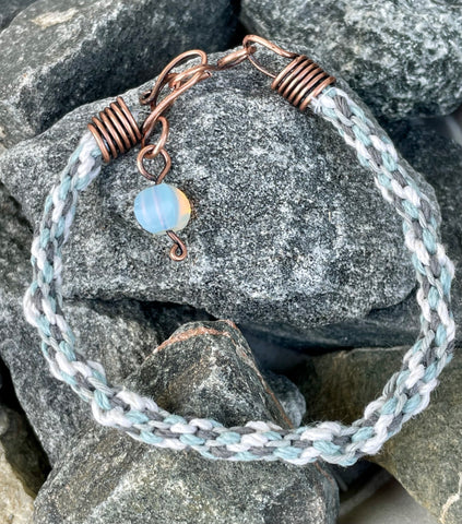 This handcrafted Kumihimo bracelet showcases a harmonious blend of blue and white hemp fibers, skillfully woven into a soft and intricate braid. The copper ends provide a warm contrast to the cool tones of the hemp, while an opalite bead dangle adds a touch of ethereal glow with its iridescent shimmer. Adjustable for a perfect fit, this bracelet is a beautiful fusion of natural textures and subtle elegance, perfect for boho and casual styles.