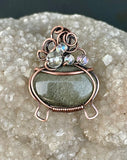 This mystical pendant features a striking Golden Sheen Obsidian gemstone, set in a handcrafted copper cauldron design. The cauldron is adorned with iridescent glass "bubbles" that shimmer with a magical glow, adding a whimsical touch to the piece. The warm copper beautifully contrasts with the dark, reflective sheen of the Obsidian, making this pendant a perfect accessory for lovers of mystical and witchy themes. A truly enchanting statement piece with a touch of magic