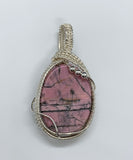 This exquisite pendant features a stunning rose-colored Rhodonite cabochon, characterized by striking black lines that create a unique and captivating design. Expertly wrapped in handwoven sterling silver (.925) and fine silver (.999), the pendant showcases exceptional craftsmanship and elegance. The soft hues of the Rhodonite combined with the lustrous silver create a beautiful contrast, making this piece a perfect addition to any jewelry collection. 