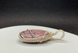 This exquisite pendant features a stunning rose-colored Rhodonite cabochon, characterized by striking black lines that create a unique and captivating design. Expertly wrapped in handwoven sterling silver (.925) and fine silver (.999), the pendant showcases exceptional craftsmanship and elegance. The soft hues of the Rhodonite combined with the lustrous silver create a beautiful contrast, making this piece a perfect addition to any jewelry collection. 