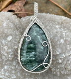 This breathtaking pendant showcases a large Seraphinite gemstone, renowned for its deep green color and feather-like patterns. The stone is intricately wrapped in Argentium Silver, known for its brilliance and tarnish resistance, creating an elegant contrast that highlights the natural beauty of the Seraphinite. Perfect for those who appreciate unique, nature-inspired jewelry, this pendant is a timeless statement piece with a graceful and ethereal charm.