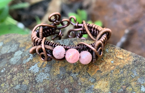 This unique adjustable ring features intricate curls, swirls, and woven copper wire accented by beautiful pink Rhodochrosite beads, creating a warm and feminine statement piece.
