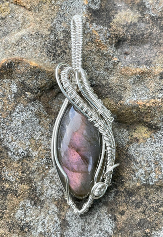 This eye-catching pendant showcases a stunning Labradorite stone with a vibrant purple and pink flash, creating a magical play of color. Wrapped in intricately handwoven Argentium Silver, known for its tarnish resistance and brilliance, this pendant exudes elegance and charm. Perfect for those who love unique, mystical gemstones, this piece adds a radiant touch to any collection.