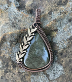 This stunning pendant features a captivating Golden Sheen Obsidian gemstone, known for its shimmering, reflective surface. Wrapped in handwoven copper, the pendant is beautifully accented with brass leaves, adding an earthy, elegant touch to the design. The warm tones of copper and brass perfectly complement the glowing Obsidian, making this a unique and eye-catching piece for anyone who loves nature-inspired, handcrafted jewelry.
