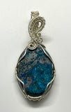 This exquisite pendant showcases a striking blend of deep blue Shattuckite and shimmering metallic Tenorite, elegantly wrapped in Argentium silver wire. The intricate wirework enhances the stone’s natural beauty while providing a sleek, modern touch. With its rich hues and artisan craftsmanship, this unique pendant is a perfect statement piece for those who appreciate rare gemstones and handcrafted elegance.