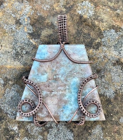 Make a bold statement with this stunning wire-wrapped pendant featuring a large lazulite gemstone in soft hues of light blue and tan. Expertly handcrafted with warm copper wire, this piece highlights the natural beauty of lazulite's intricate patterns. The rich copper tones perfectly complement the stone’s serene colors, creating a timeless accessory that's ideal for nature lovers and bohemian styles alike.