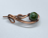 This exquisite shawl pin is crafted from hand-shaped heavy gauge copper, skillfully combined with Copper Ore Jasper. The robust copper provides a sturdy frame, while the Copper Ore Jasper adds a natural and unique touch. The design elegantly secures your shawl or scarf, merging functionality with beauty. This piece stands out with its earthy tones and artisanal craftsmanship, perfect for adding a distinctive flair to your outfit.