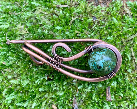 This exquisite shawl pin is crafted from hand-shaped heavy gauge copper, skillfully combined with Copper Ore Jasper. The robust copper provides a sturdy frame, while the Copper Ore Jasper adds a natural and unique touch. The design elegantly secures your shawl or scarf, merging functionality with beauty. This piece stands out with its earthy tones and artisanal craftsmanship, perfect for adding a distinctive flair to your outfit.