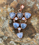 This Copper Pendant featuring 6 Blue Dumortierite Cabochons and 3 Faceted Rainbow Moonstones is a striking and intricate piece of jewelry. The pendant showcases a sophisticated blend of blue dumortierite and rainbow moonstone, all elegantly set in copper. This pendant embodies a harmonious combination of deep, rich colors and vibrant, iridescent highlights.