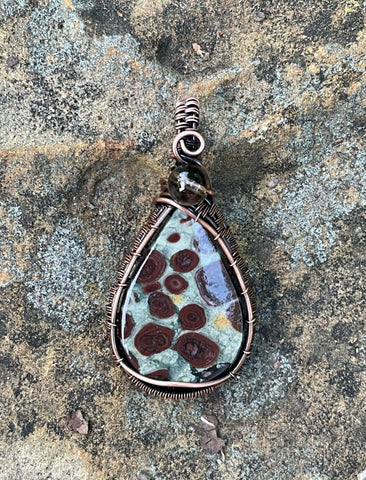 This handcrafted wire-wrapped copper pendant features a stunning Asteroid Jasper, known for its unique patterns and cosmic-like appearance, paired with a faceted Smoky Quartz accent. The warm copper wire enhances the earthy tones of the stones.