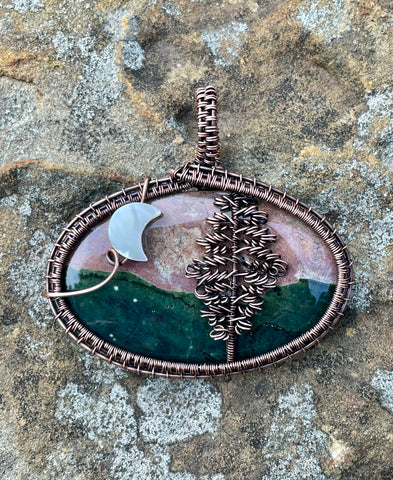 This Ocean Jasper Pendant is a beautifully handcrafted jewelry piece wrapped in Copper, featuring a striking Tree design alongside a delicate Moon accent. The pendant's centerpiece is a naturally vibrant Ocean Jasper stone, showcasing a range of earthy tones that resemble the serene beauty of nature.